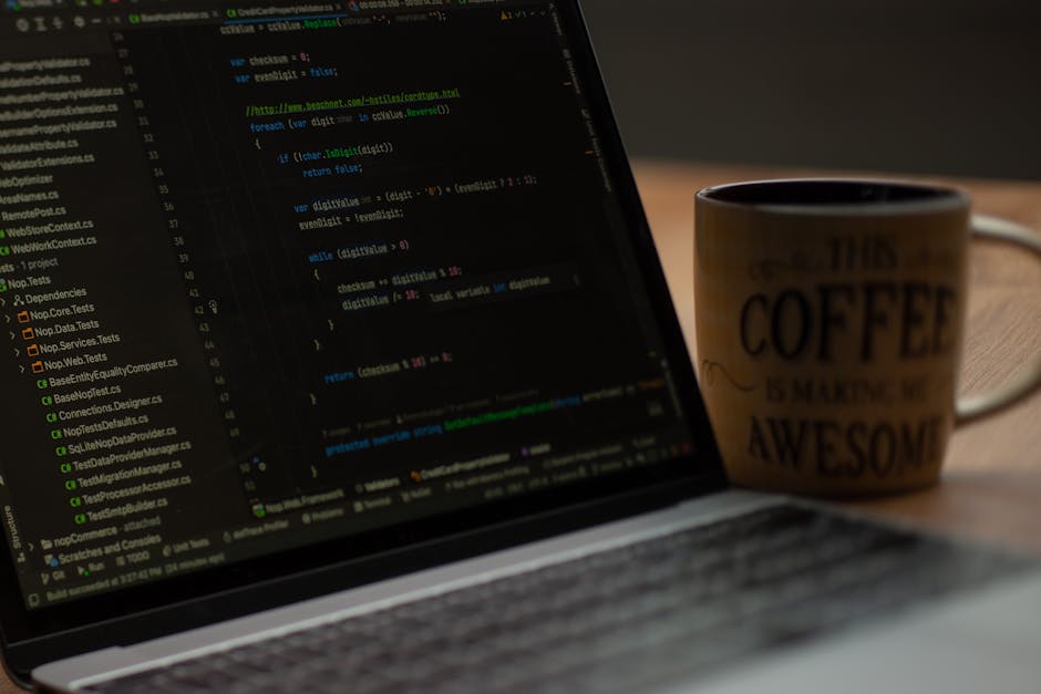 A laptop displaying lines of programming code on its screen, placed on a wooden desk alongside a ceramic coffee mug with the phrase 'This coffee is making me awesome' printed on it. The workspace feat